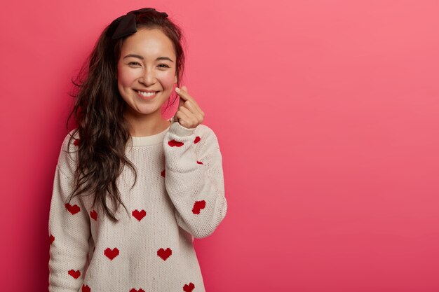 Romantic Chinese woman shows korean heart sign with two fingers crossed, smiles joyfully and confesses in love, expresses affection, wears sweater with heart print, isolated on pink studio wall