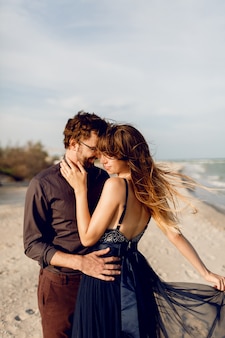 Abbraccio romantico delle coppie sulla spiaggia di sera vicino all'oceano. donna elegante in vestito blu che abbraccia il suo ragazzo con tenerezza.