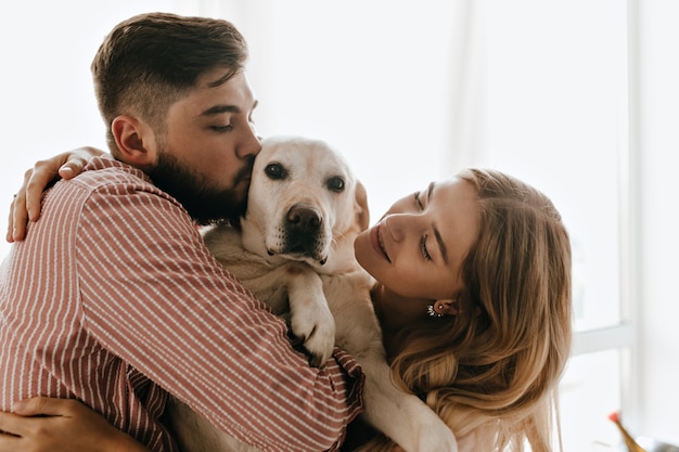 Foto gratuita coppia romantica di buon umore gioca e abbraccia il cane bianco. l'uomo bacia il suo labrador contro la finestra.