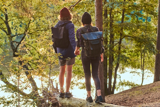 Romantic couple is standing near river in green bright forest and looking foward. They have backpacks and hats.