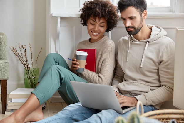 Romantic couple sit closely on floor, focused in laptop computer, watch interesting film online, enjoy aromatic coffee, being in good mood, enjoy wireless internet connection, have spare time
