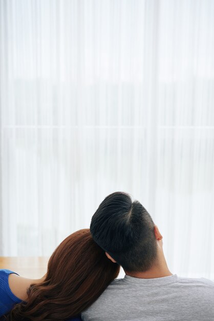 Romantic couple sitting together on couch at home in front of window and touching heads