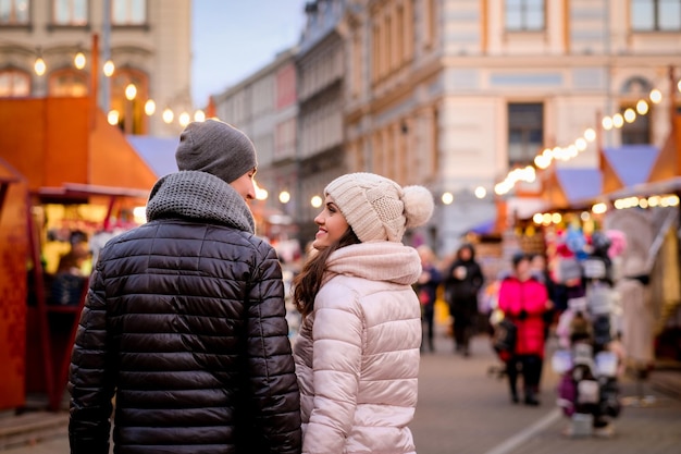Free photo romantic couple wearing winter clothes hugging while standing in evening street with christmas fair