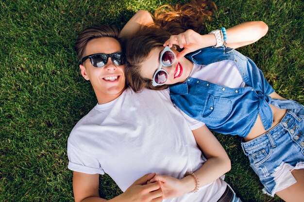 Romantic couple of young people in sunglasses is lying on grass in park. Girl with long curly hair is lying on shoulder of handsome guy in white T-shirt.  View from above.