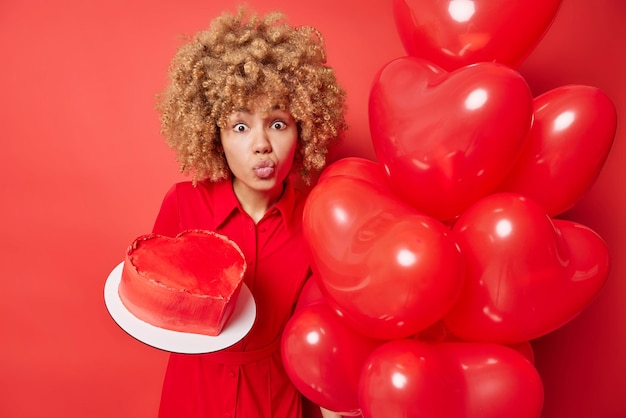 Romantic curly haired woman keeps lipd folded holds delicious cake in form of heart and inflated balloons wears dress isolated over vivid red background People holidays celebration concept