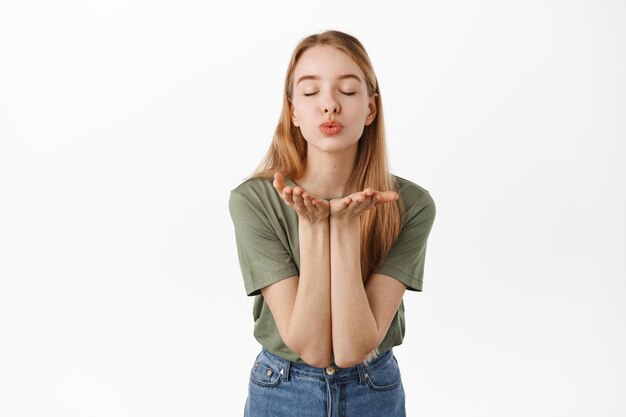 Romantic cute girlfriend sending air kiss at camera with closed eyes, puckered lips and hands near mouth, standing over white background