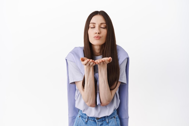 Romantic cute woman blowing air kiss at camera, close eyes and send mwah, standing against white background