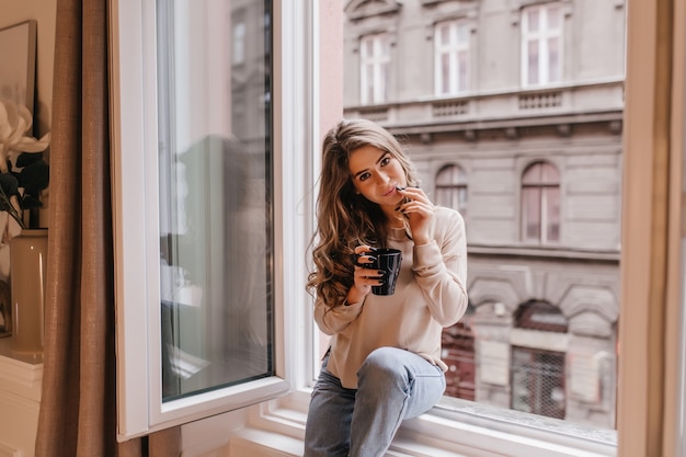 Romantic dark-haired woman spending time at home and drinking latte on sill