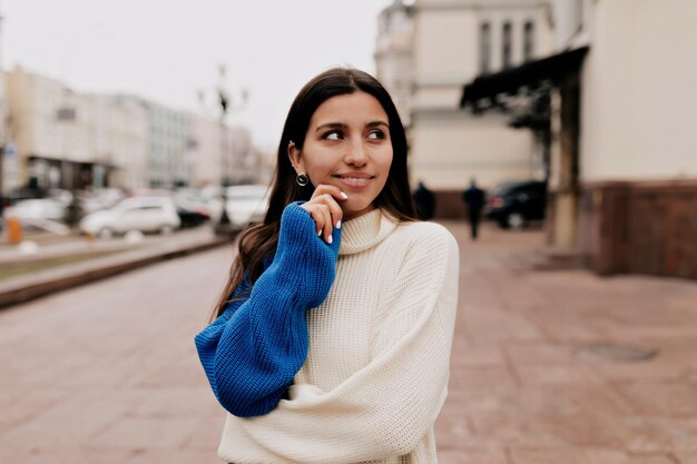 Romantic European woman with lovely smile and dark hair is wearing spring bright knitted sweater is looking up and holding hand on the lips