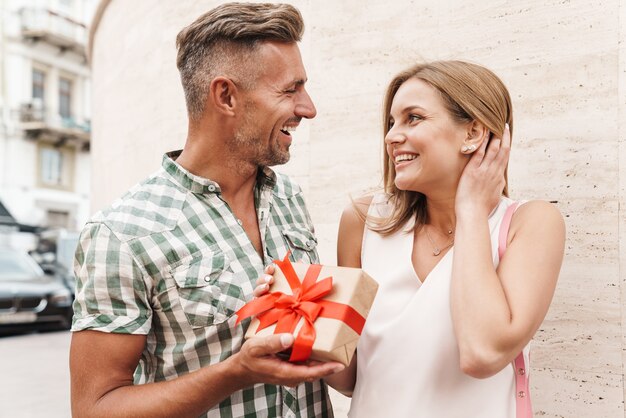 街の通りの壁に立っている間、笑顔でプレゼントボックスを一緒に保持している夏服のロマンチックな興奮したカップル