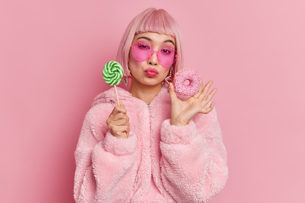 로맨틱 세련된 핑크 머리 글래머 여성 모델은 달콤한 이빨이 롤리팝과 따뜻한 모피 코트 유행 심장 모양의 선글라스를 입은 유약 도넛을 보유하고 있습니다.