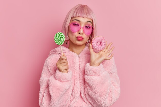 Romantic fashionable pink haired glamour female model has sweet tooth holds lollipop and glazed doughnut dressed in warm fur coat trendy heart shaped sunglasses 