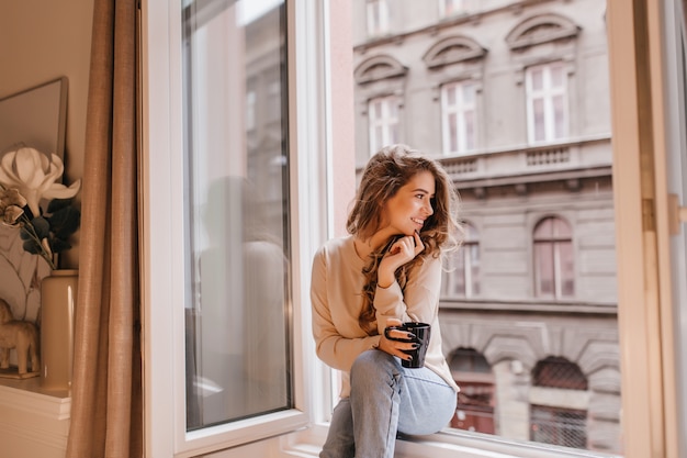 Romantic female model in good mood looking at street, sitting on sill