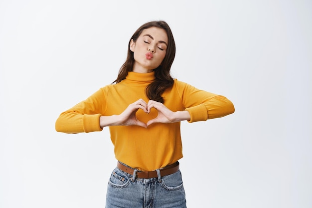 Romantic girl fall in love, showing heart gesture with closed eyes and kissing lips, standing on white