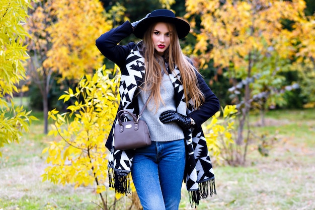 Romantic girl in glasses holding hat and posing with kissing face expression standing in the middle of park. Outdoor portrait of cute young woman with trendy scarf having fun during walk in forest.