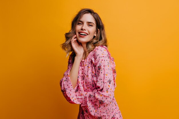 Romantic girl in pink blouse posing with sincere smile. Indoor photo of pretty curly woman having fun on yellow wall.