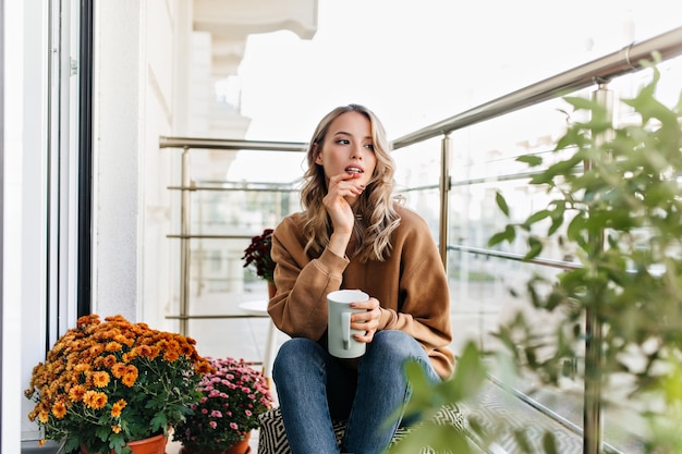 Романтичная девушка сидит возле цветов с чашкой кофе. Беззаботная молодая женщина думает о чем-то.