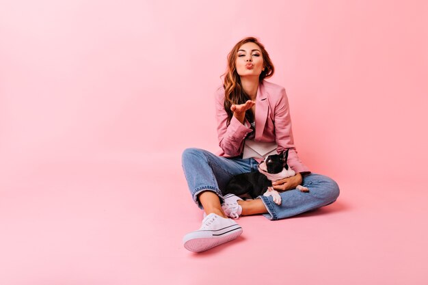 Romantic girl in stylish outfit sending air kiss during portraitshoot with her dog. Indoor portrait of blithesome ginger woman having fun with pet.