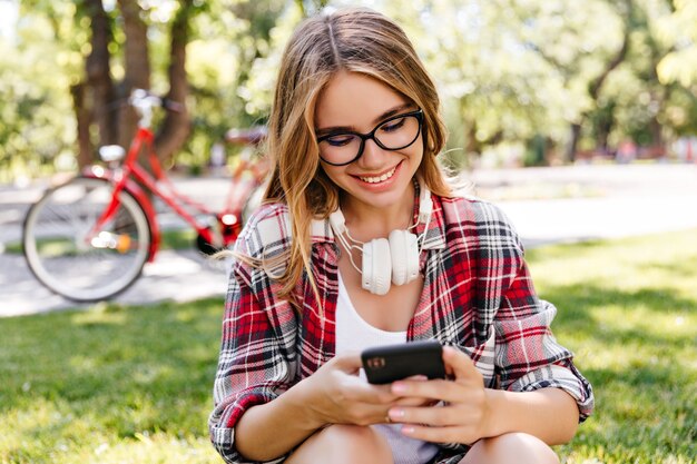 Romantic girl texting message while resting in beautiful park. Outdoor photo of cheerful blonde woman sitting on grass with smartphone.