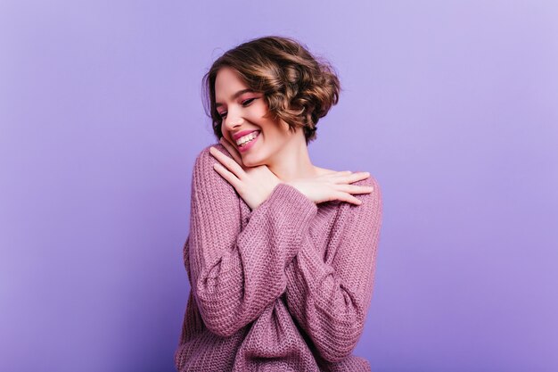 Романтичная девушка с элегантным макияжем наслаждается фотосессией в помещении в уютной одежде. Симпатичная брюнетка молодая женская модель в вязаном свитере, стоящем с улыбкой на фиолетовой стене.