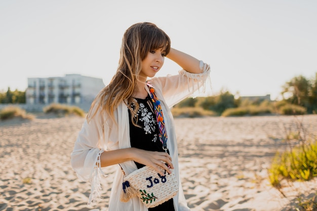 Romantic girl with long  brunette hair walking on the beach.   Bohemian style, straw bag , bright make up. Tropical mood. Sunset colors.