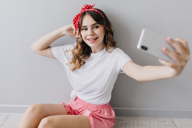 Romantic girl with red ribbon in hair taking picture of herself and laughing. Indoor shot of inspired woman in white t-shirt making selfie.