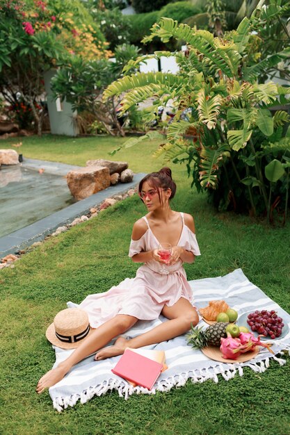 Romantic graceful Asian model sitting on blanket, drinking wine and enjoying summer picnic in tropical garden.