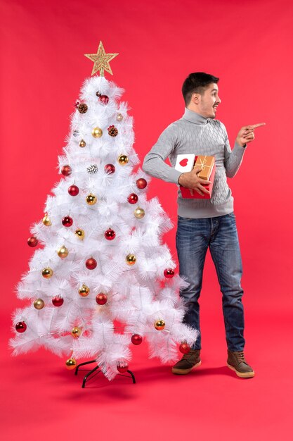 Romantic handsome adult in a gray blouse standing near the decorated white Christmas tree