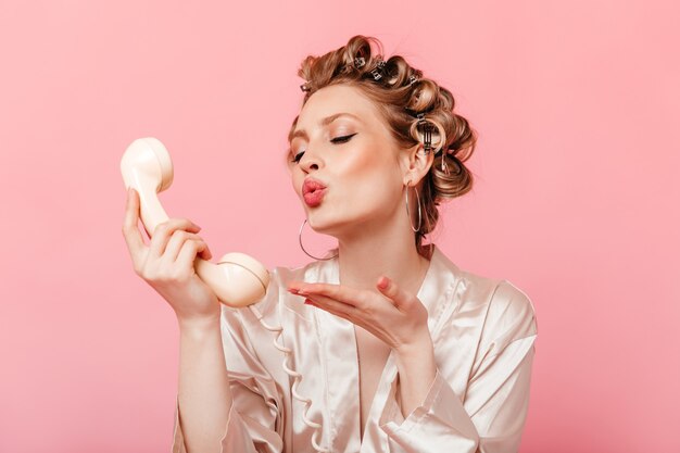 Romantic housewife in hair curlers on her head and light robe sends kiss on phone