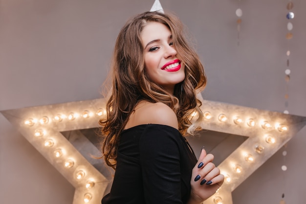 Romantic laughing woman with bright makeup having fun on indoor photoshoot on dark wall. Pleased white girl with trendy hairstyle relaxing .