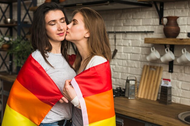 Romantic lesbian couple wrapped in rainbow flag standing in the kitchen