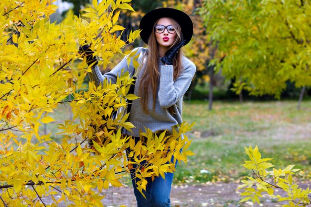 Romantic long-haired girl posing with kiss face expression while walking in autumn park. Outdoor portrait of elegant european young woman in jeans and hat standing beside yellow bush.