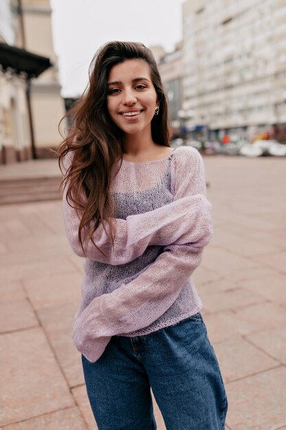 Romantic lovely woman with long wavy dark hair is wearing tender spring thin sweater is posing at camera and smiling