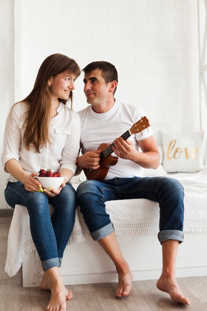 Романтичный человек играет на гавайской гитаре и смотрит на свою жену, держащую миску с клубникой