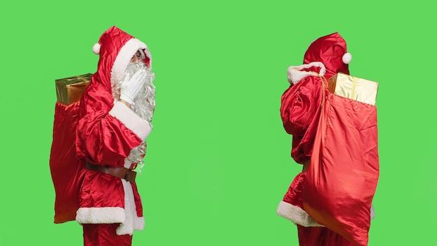 Foto gratuita uomo romantico in costume da babbo natale che dà baci d'aria, carino mentre porta il tradizionale sacco festivo con regali. il dolce santo nick fa un gesto provocante stando in piedi sullo sfondo con schermo verde.