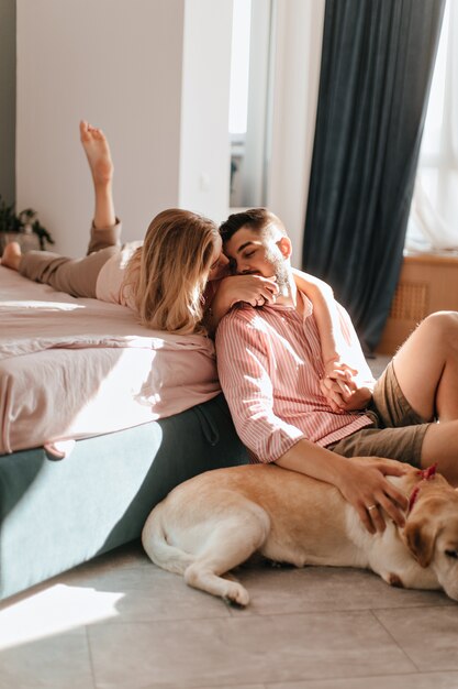 Romantic married couple hugging. Man strokes labrador and chats with his beloved woman lying on bed.