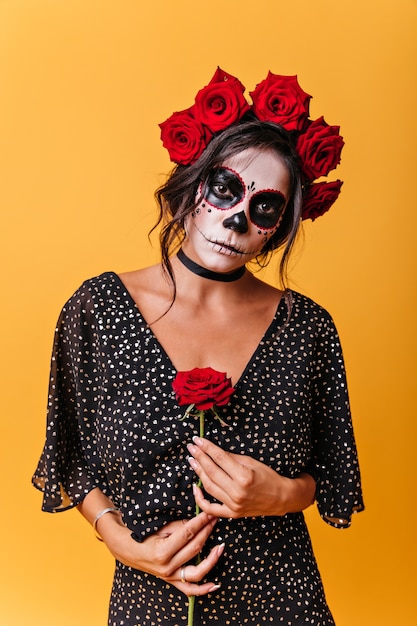 ロマンチックなメキシコ人は優しくバラを保持します。彼女の髪とハロウィーンの化粧に花を持つ謎の女性の肖像画。