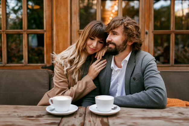 Romantic pensive woman with long wavy hairs hugging her husband with beard. Elegant couple sitting in cafe with hot cappuccino.