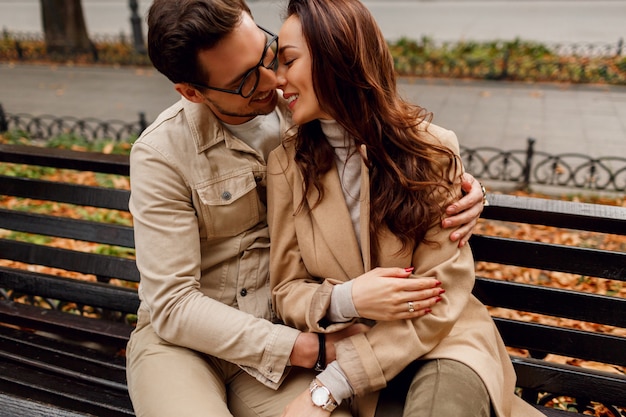 秋の公園でベンチにハグ、キスの愛の若い美しいカップルのロマンチックな肖像画。スタイリッシュなベージュのコートを着ています。