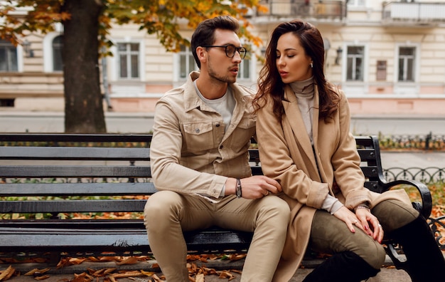 Romantic portrait of  young beautiful couple  in love hugging and kissing  on bench in autumn park. Wearing stylish beige coat.