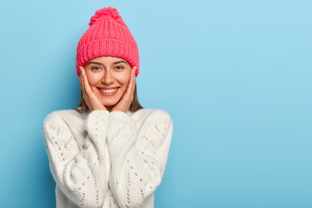 Romantic positive young european woman smiles gently, has white perfect teeth, touches both cheeks, has friendly look, wears pink hat with pompon and white sweater