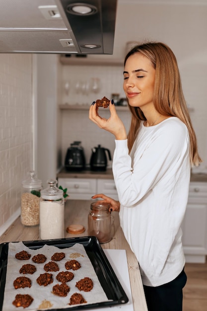 Romantic pretty girl with long hair wearing white shirt is tasting her cookies with closed eyes Charming girl at kitchen with cookies