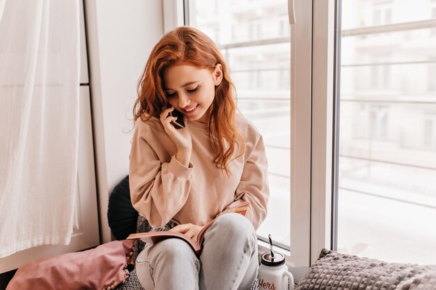 Romantic red-haired girl reading book beside window. Shy european young woman talking on phone.