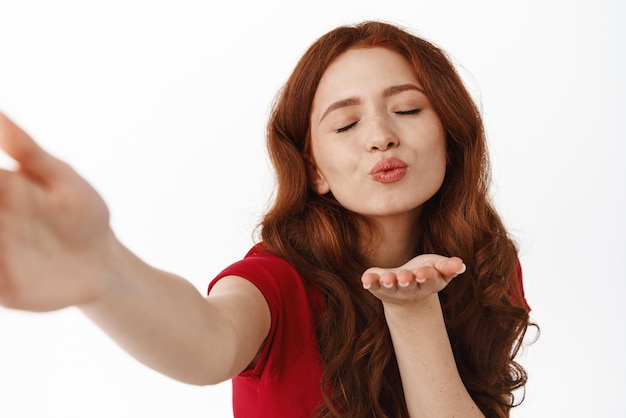 Romantic redhead woman sending air kiss with cute flirty face taking selfie or video calling lover flirty and coquettish goodbye standing against white background