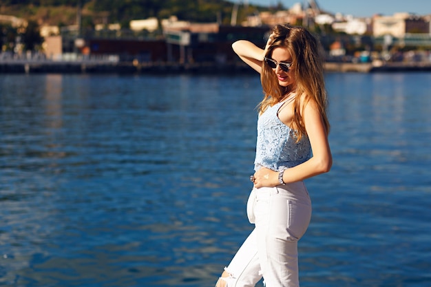 Romantic sea view portrait of sensual blonde woman,trendy summer outfit, pastel colors, travel alone, vacation, white denim, sunglasses.