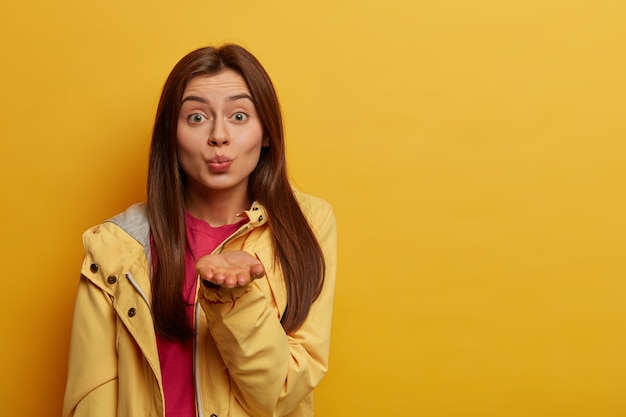 Romantic sensual woman keeps lips rounded, blows air kiss at camera, expresses affection, has tender look, wears yellow anorak, stands indoor, copy space area. People