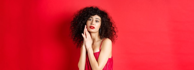 Free photo romantic sensual woman with curly hair wearing evening dress posing seductive on red background