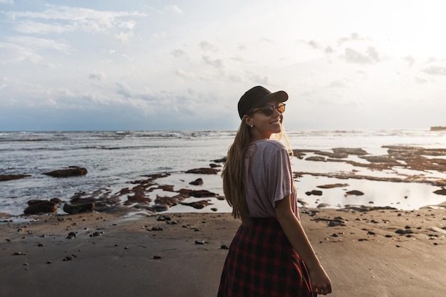 Romantic shot of beautiful carefree woman backpacker travel around world trip to Indonesia Bali look back to smile camera as enjoying relaxing view on black sandy volcanic beach and ocean shore