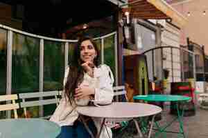 Free photo romantic shy girl with long hair wearing white coat sitting on french cafe outdoors and waiting for date