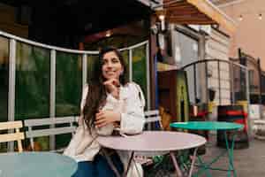 Бесплатное фото Романтичная застенчивая девушка с длинными волосами в белом халате сидит во французском кафе на открытом воздухе и ждет свидания
